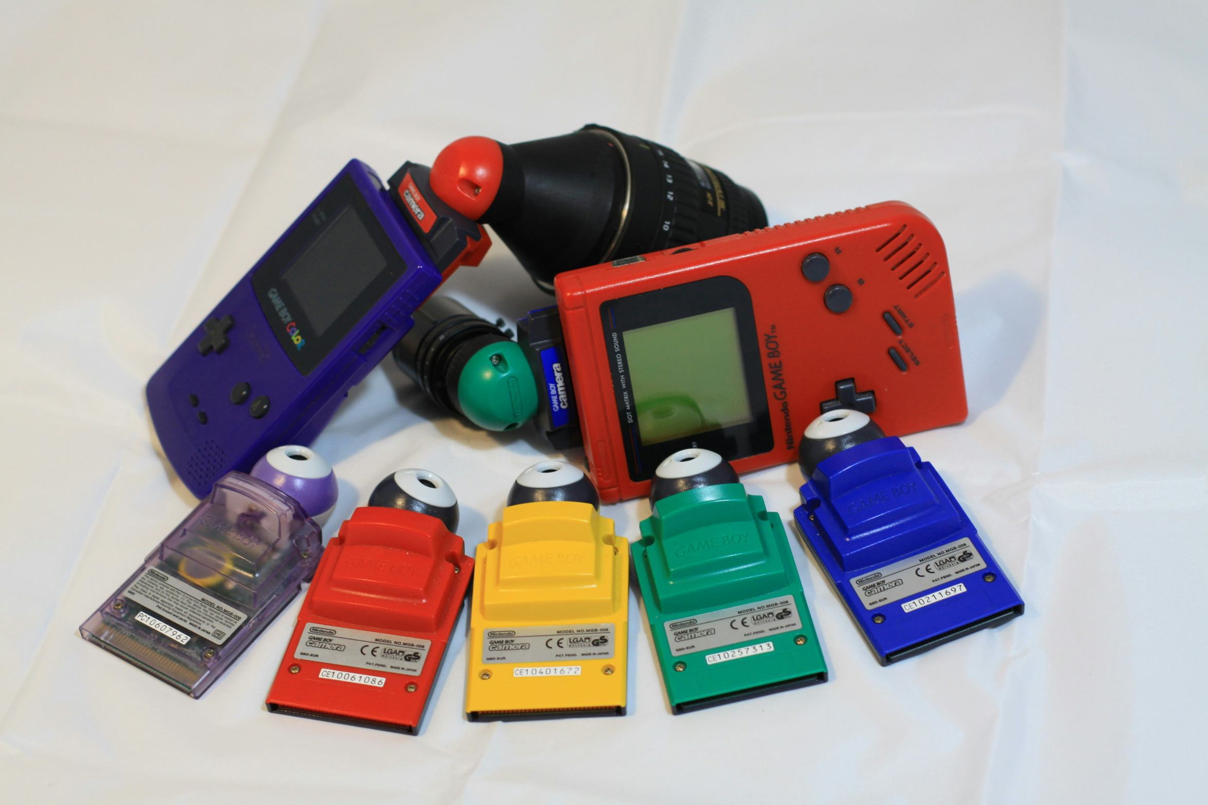 A family of different colored Game Boy Cameras with Game Boys and DIY camera lens attachments.