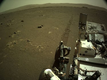 Perseverance rover seen in foreground on the surface of Mars, with tire marks trailing behind it