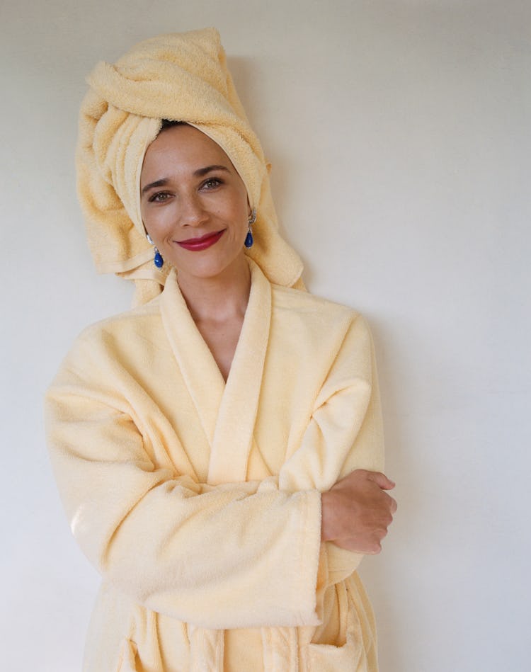 Rashida Jones smiling in a yellow bathrobe and hair tucked in the yellow towel with blue earrings