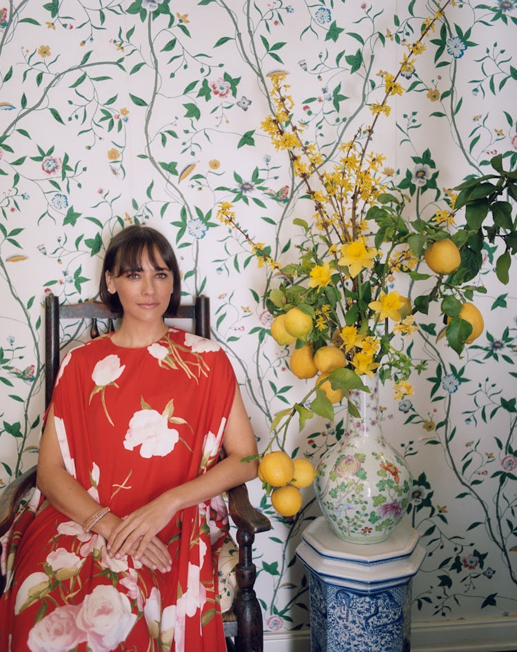 Rashida Jones sitting in front of the floral wallpaper, vase, and vase holder in a vibrant floral dr...