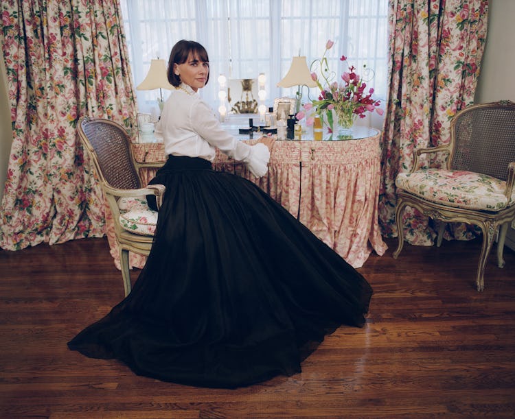 Rashida Jones sitting in front of the vintage table and floral curtains in a black maxi skirt & whit...