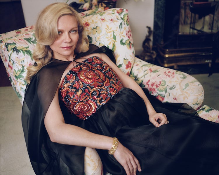Kirsten Dunst lying on a floral armchair in black dress with a glitter corset and black cape
