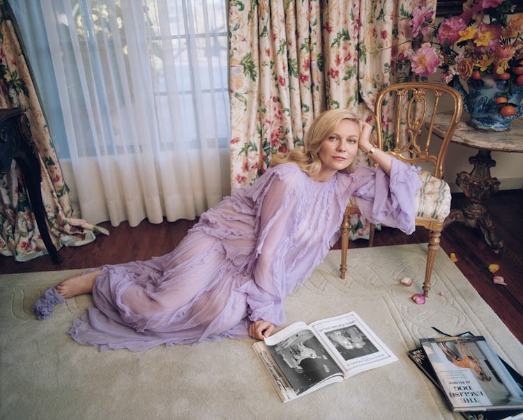 Kirsten Dunst sitting in a sheer lilac dress on the floor with her hand on a vintage chair and readi...