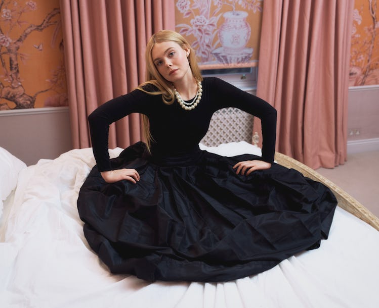 Elle Fanning sitting on the bed in a black maxi skirt & black top with white pearls