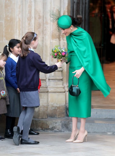 Meghan Markle smiling in a green dress and cape and a green hat receiving flowers from a group of ch...