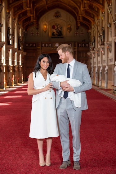 Meghan Markle in a white dress and Prince Harry in a grey suit standing and posing with their newbor...