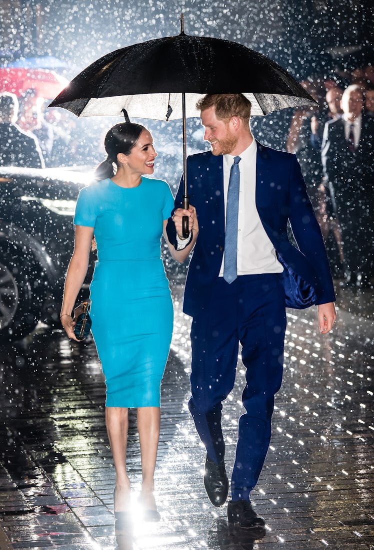 Meghan Markle in a blue dress and Prince Harry in a navy suit walking with an umbrella while it's ra...