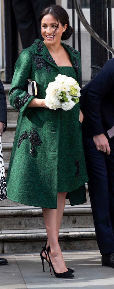 Meghan Markle in a green dress and coat walking and smiling white holding white roses