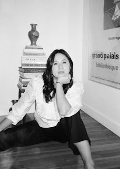 A black and white photo of Chelsea Mak, posing on the floor with a books stacked up behind her 