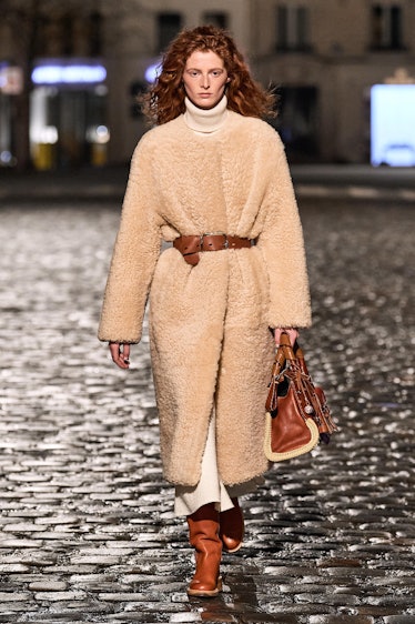 A model walking the runway in a beige jacket with a brown belt with matching shoes and bag by Chloé