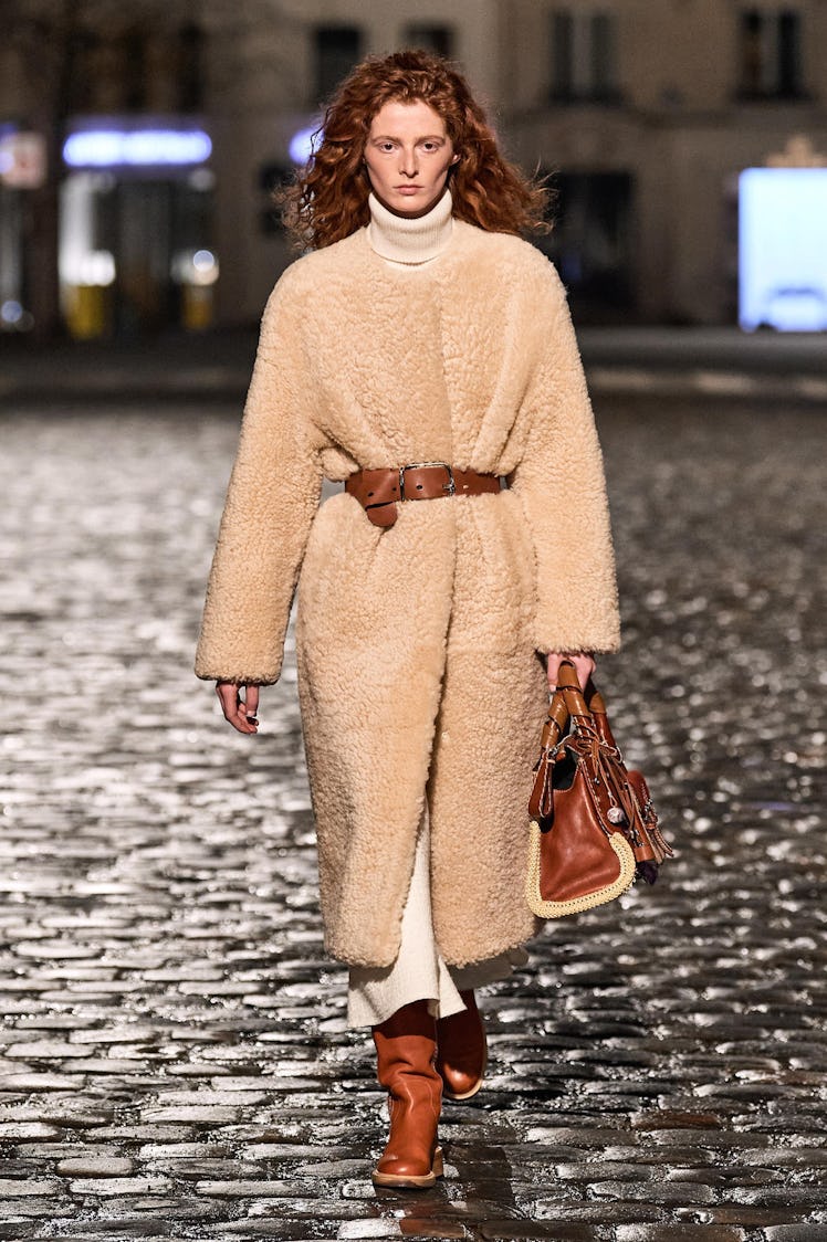 A model walking the runway in a beige jacket with a brown belt with matching shoes and bag by Chloé