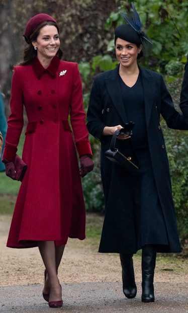 Meghan Markle in a black dress and coat and Kate Middleton in a red coat walking and smiling