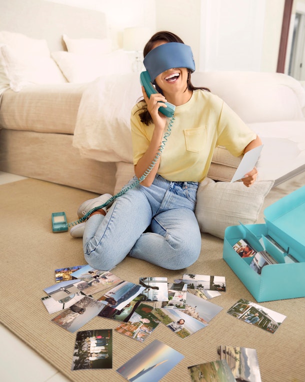 A young woman sits on her living room floor with travel photos while wearing a sleep mask from Away's Travel Well collection.