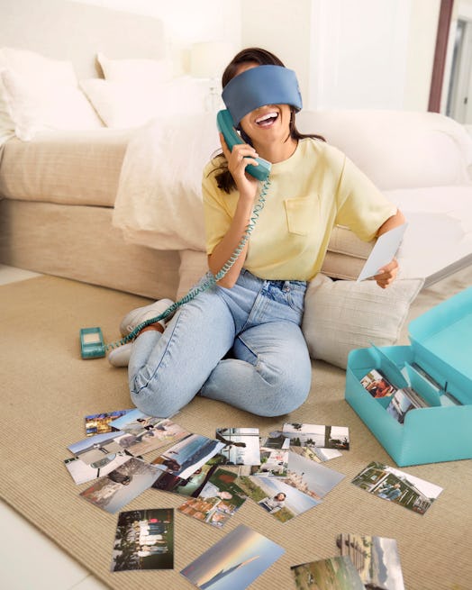A young woman sits on her living room floor with travel photos while wearing a sleep mask from Away'...