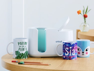 A Cricut Mug Press sits on a table with one-of-a-kind custom DIY mugs. 