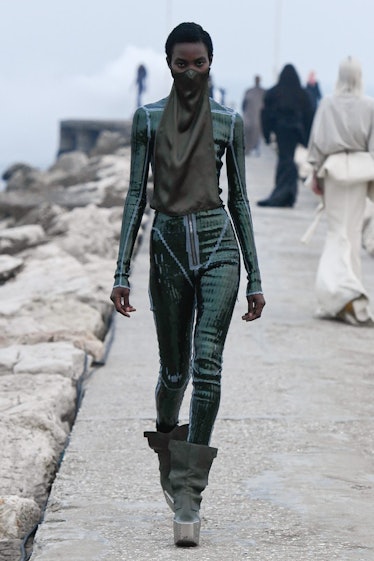 A model in a Rick Owens green shimmery jumpsuit and green boots with a face mask that extends into a...