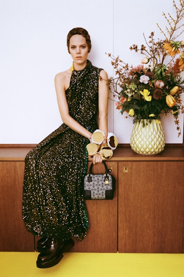 A model sitting on a cupboard in a black dress with multicolored spots on it by Loewe