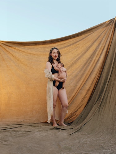 Nicole Stark, holding her baby, wearing a robe, standing in front of a backdrop with blue sky behind...