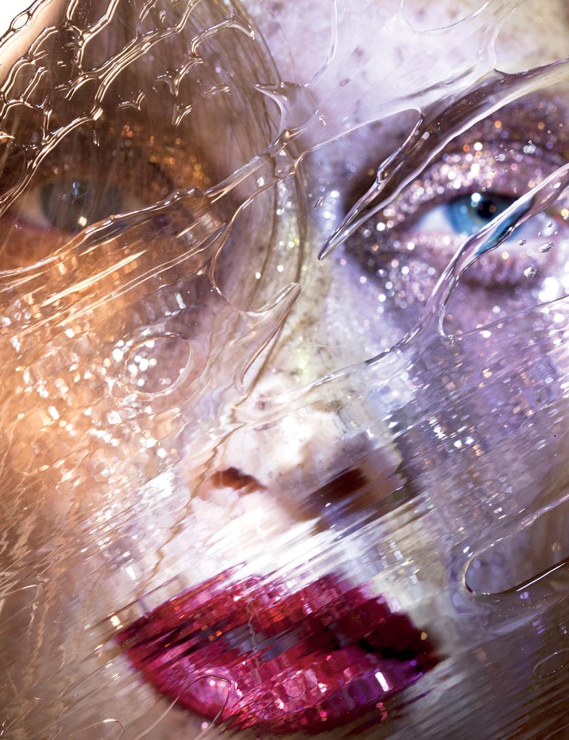 A close-up of a woman's face covered with flitter on her skin and red lips behind a water stained su...