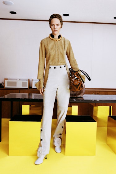 A model in a suede beige shirt, white pants and hells with a brown bag all by Loewe