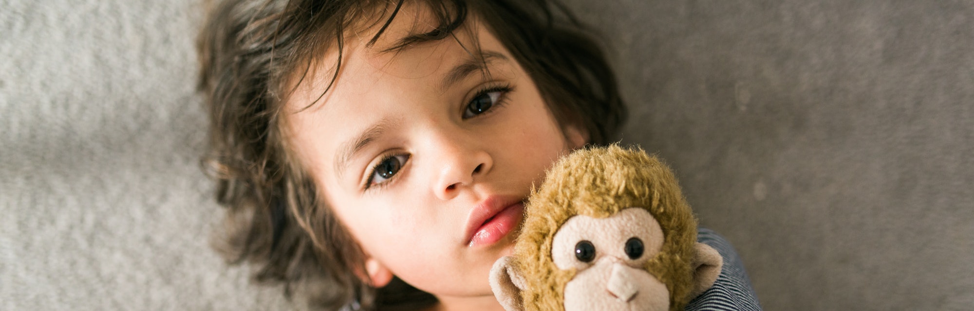 A small child looks up at the viewer, as he snuggles with his stuffed animal monkey on the floor