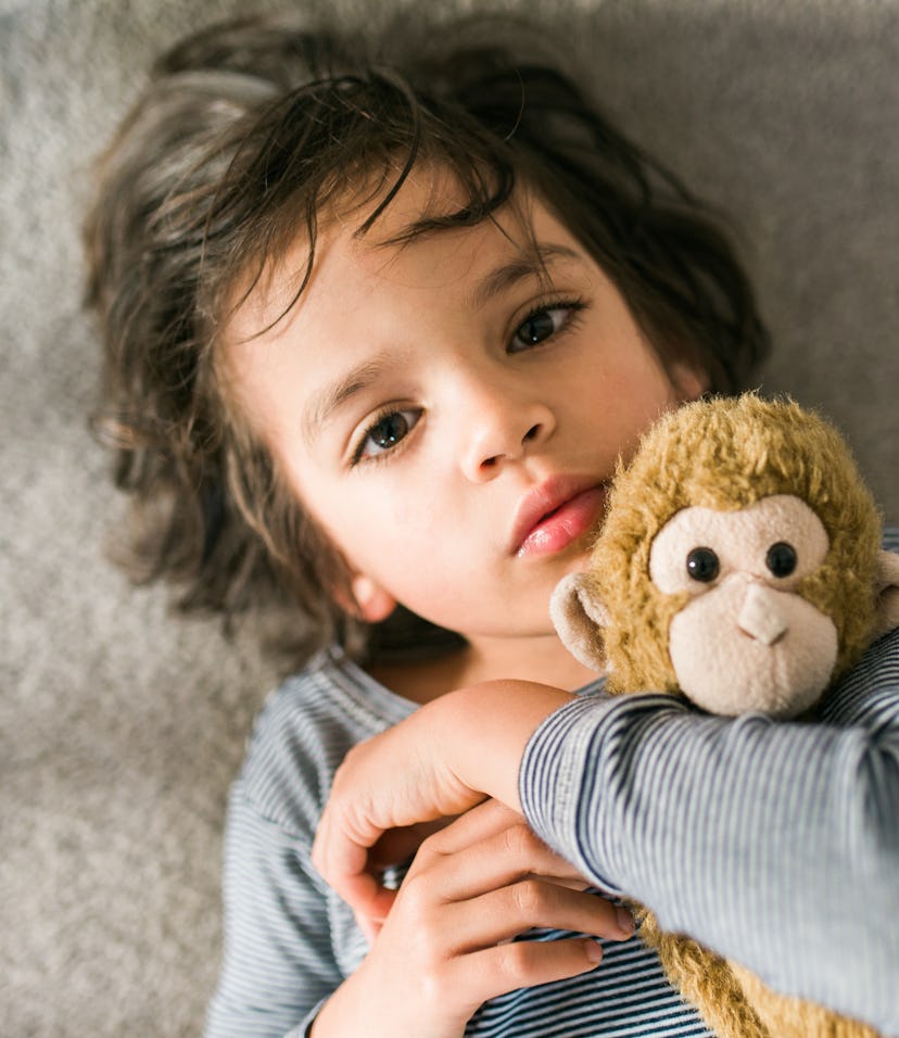 A small child looks up at the viewer, as he snuggles with his stuffed animal monkey on the floor
