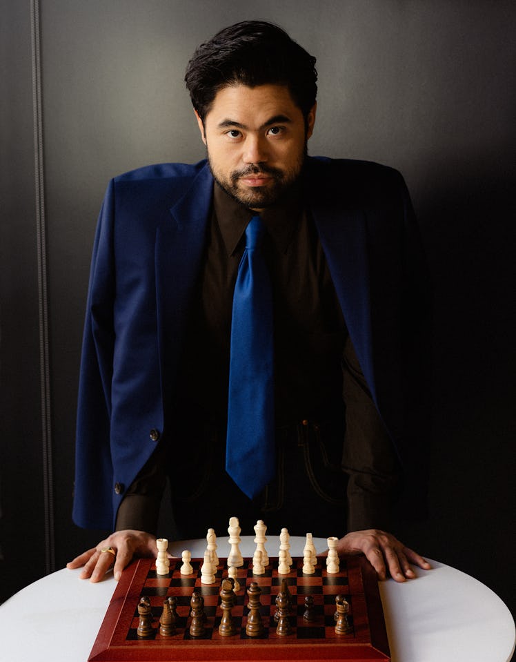 Hikaru Nakamura with a chess board in front him, wearing a black shirt with a dark blue blazer and m...