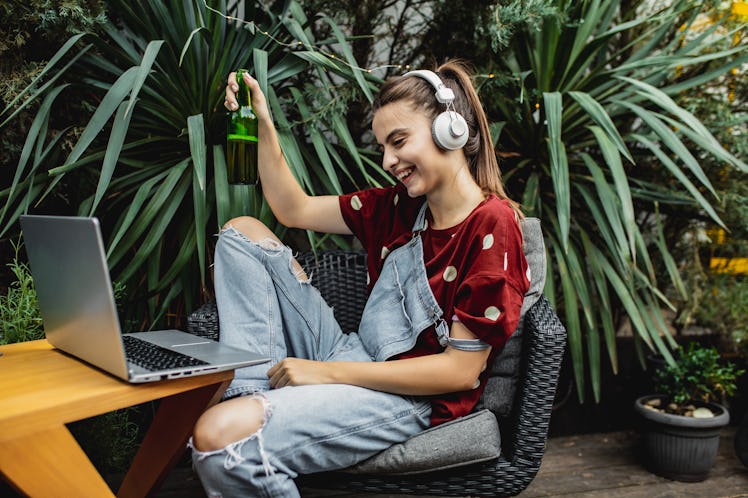 A young woman laughs while holding a beer and attending a virtual St. Patrick's Day party with games...