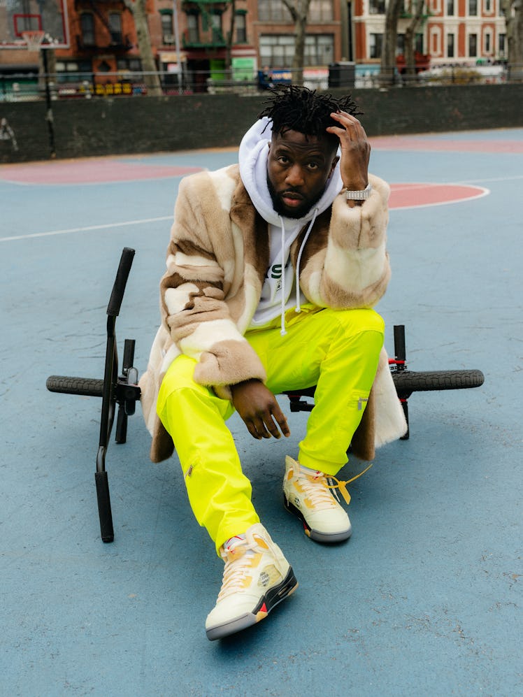 Nigel Sylvester in a faux fur coat, white sweatshirt , neon yellow pants and beige sneakers on a bas...