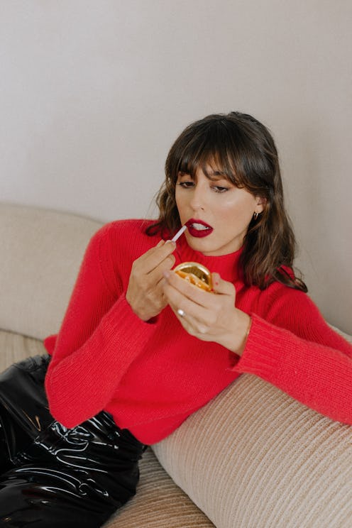 Violette_Fr in a red sweater and black vinyl pants applying burgundy lipstick from her own brand