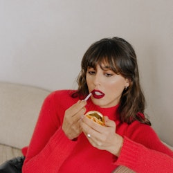Violette_Fr in a red sweater and black vinyl pants applying burgundy lipstick from her own brand