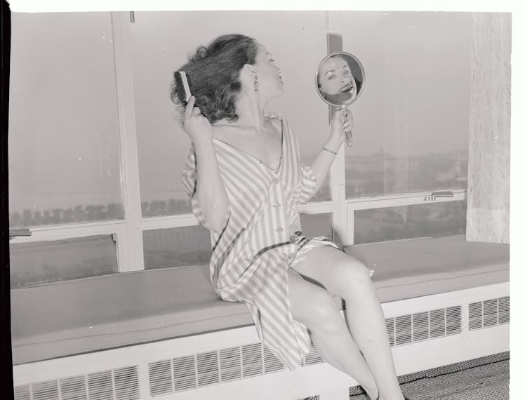 A woman brushing her hair while holding a small hand mirror, wearing a striped dress