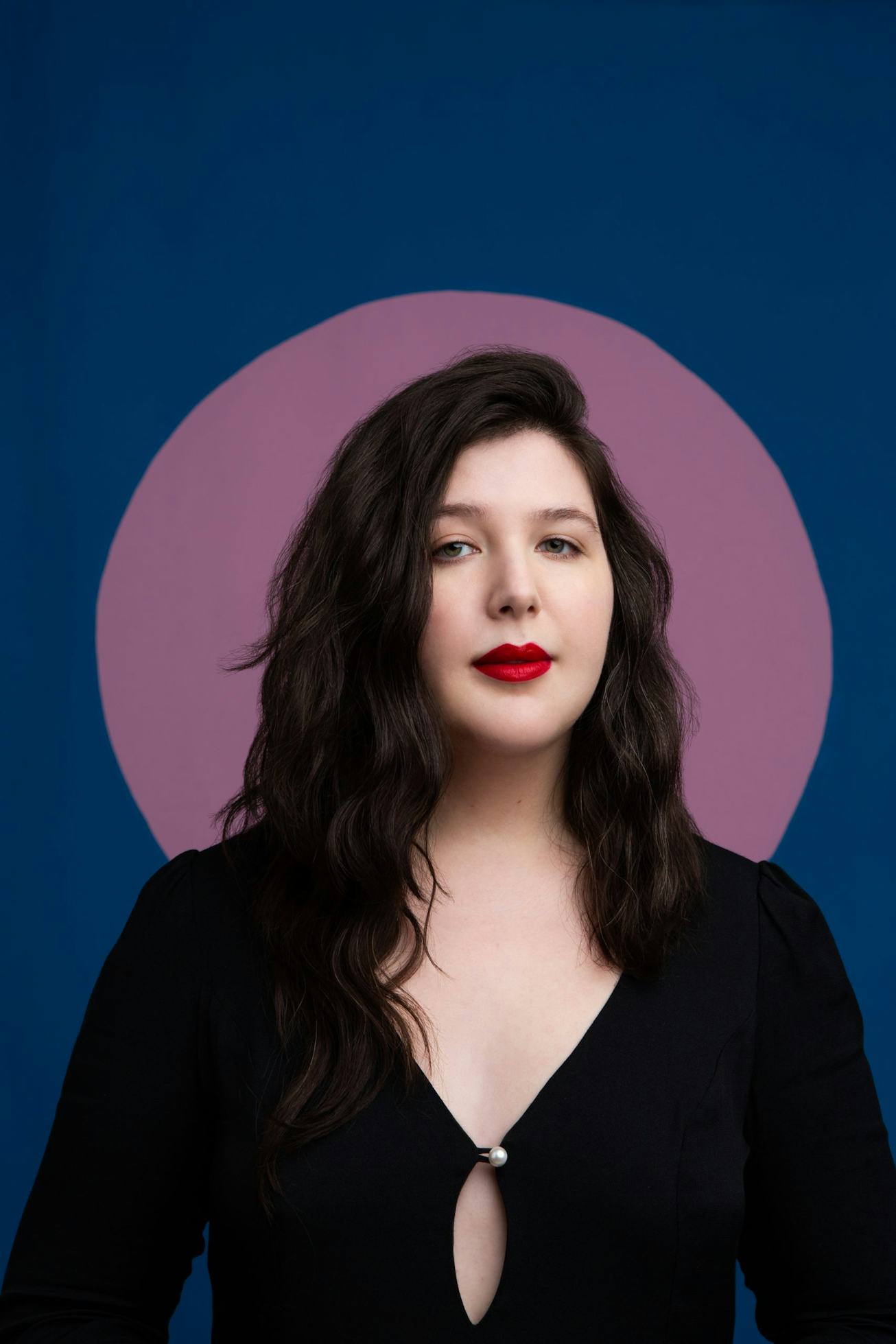 A portrait of Lucy Dacus. She stands in front of a blue background with a lavender circle framing he...