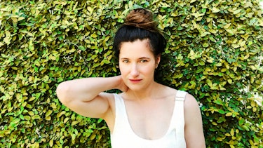 Kathryn Hahn in white overalls standing in front of a hedge, with her hair in a topknot
