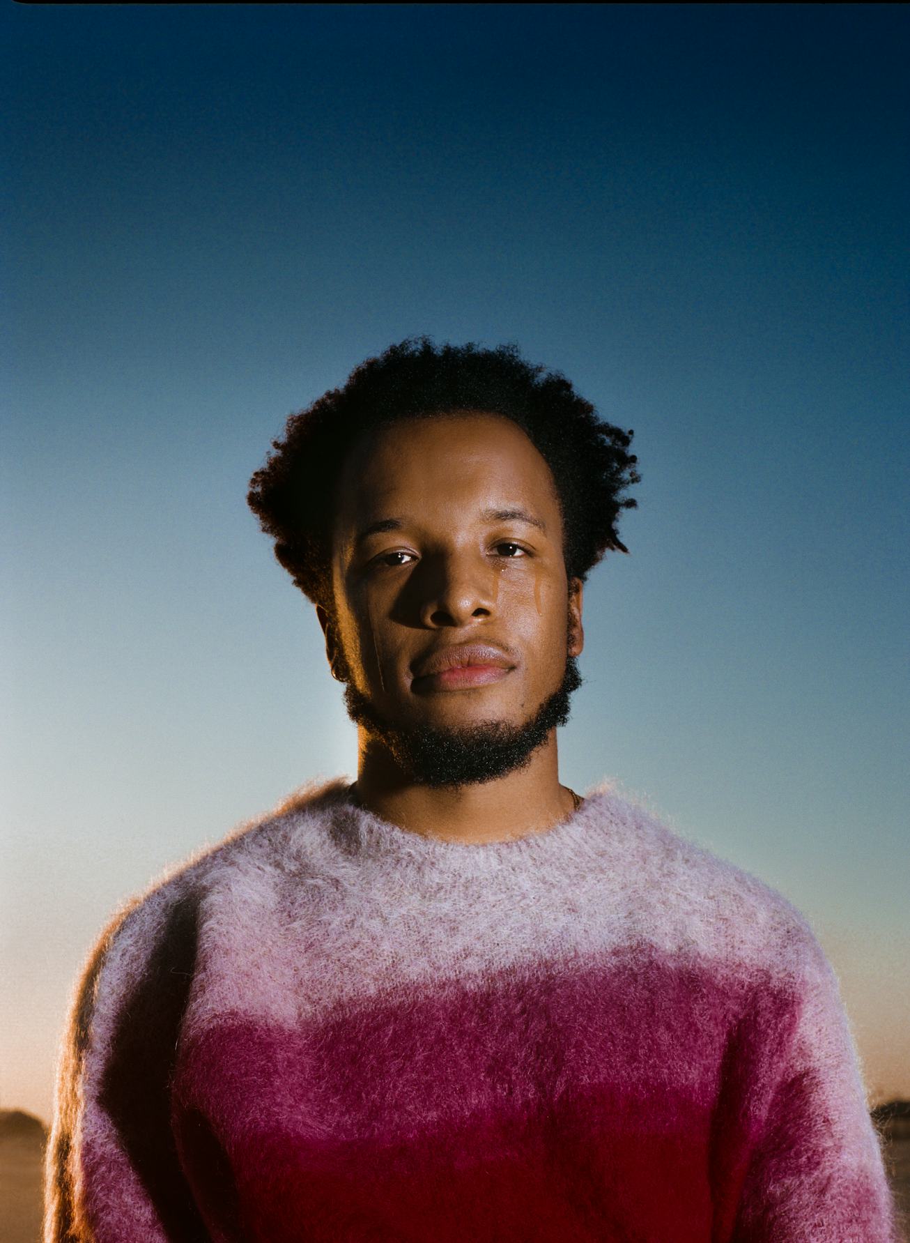 A portrait of Cautious Clay. He's in a pink sweater and standing against an ombre sunset sky.