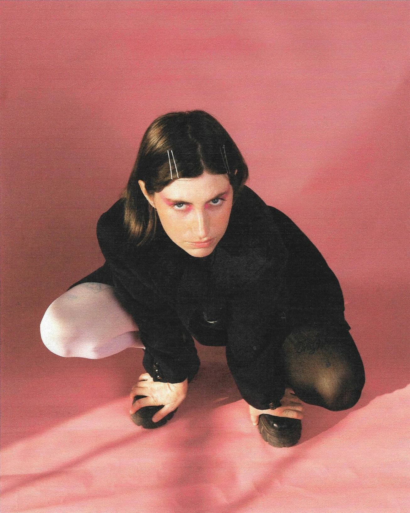 A portrait of Ian Sweet. She's kneeling against a pink background with rosy eyeshadow on, and an all...