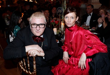 Alber Elbaz in a black suit sitting and posing next to Audrey Marnay in a red dress