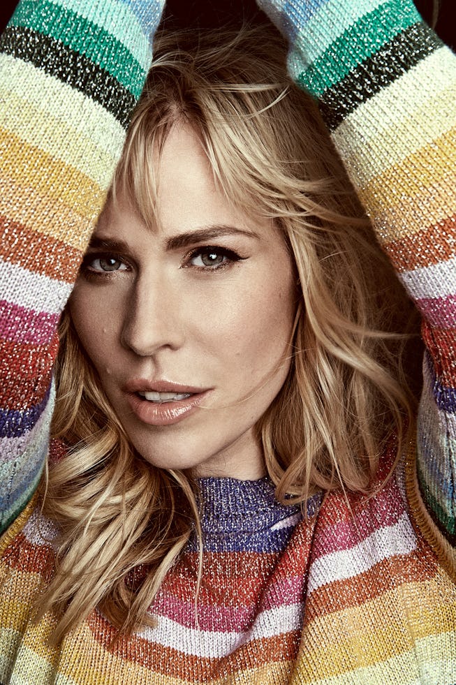 A portrait of Natasha Bedingfield. She wears a rainbow striped sweater and has her arms up framing her face.