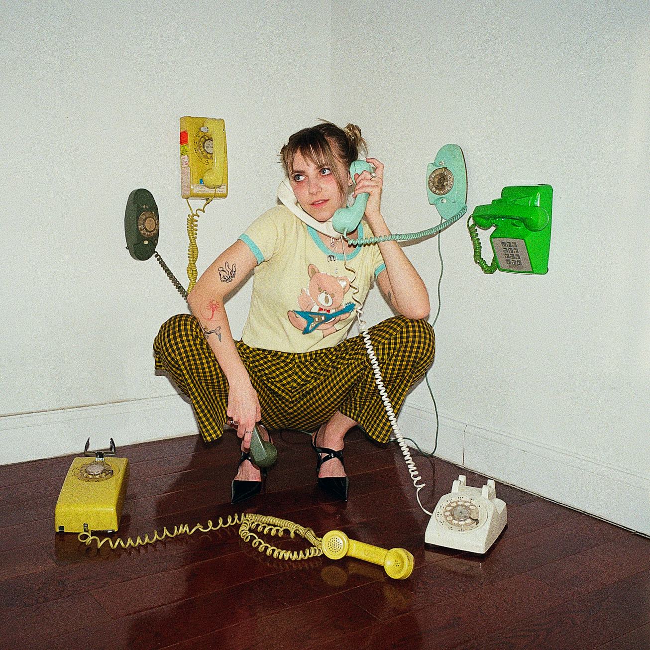 A portrait of May Rio. She's squatting surrounded by several landline phones.