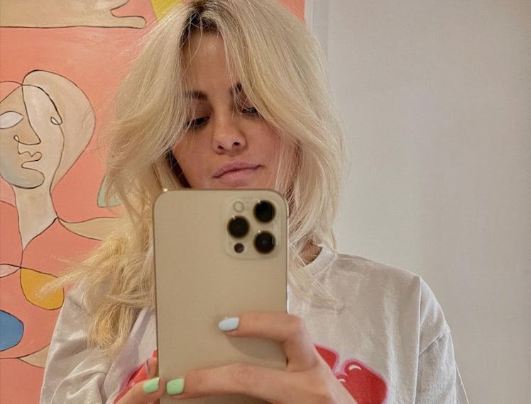 Selena Gomez with blonde hair taking a mirror selfie in a white shirt