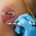 A close-up of an in-progress tattoo on a person's arm that reads, "Mom"