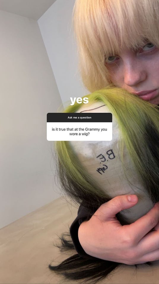 Billie Eilish with platinum blonde hair holds the black and green wig she wore to the Grammys 2021