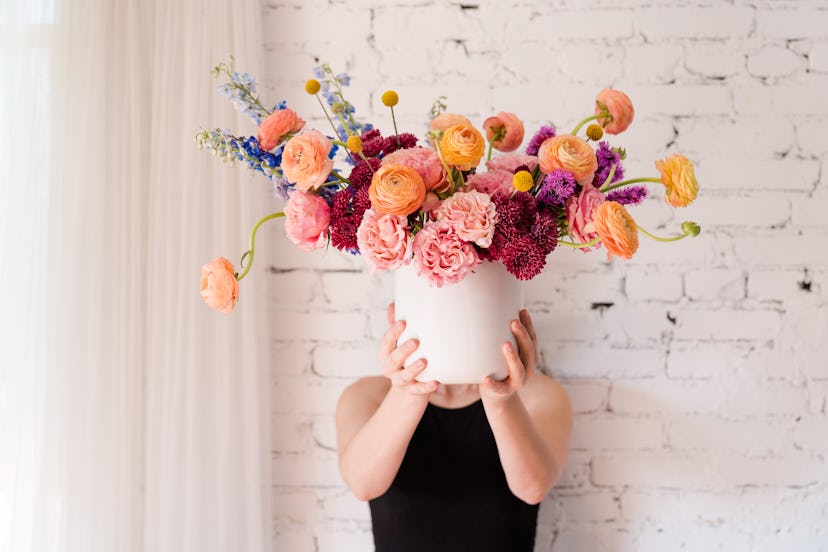 a woman holds a bouquet of flowers in a white bucket, including blue snapdragon, peach ranunculus an...