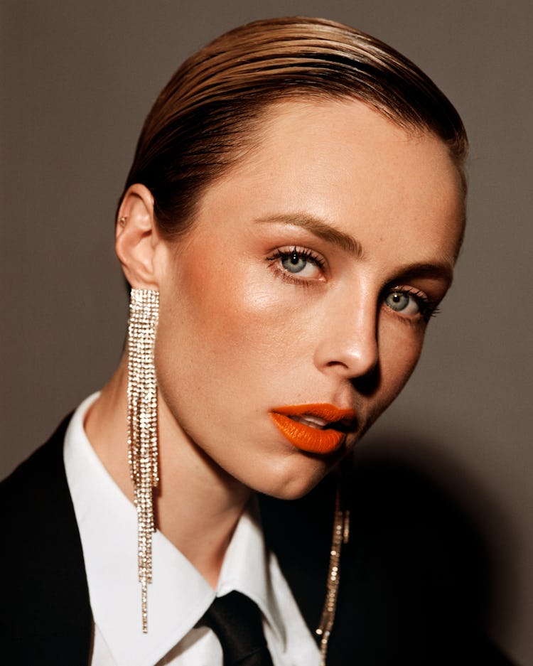 Edie Campbell in a white button-up with a black suit and tie, long dangling earrings and orange lips...