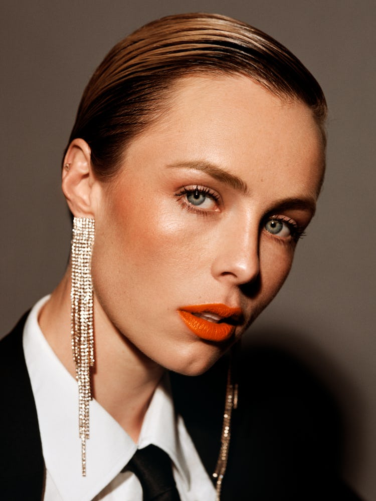 Edie Campbell in a white button-up with a black suit and tie, long dangling earrings and orange lips...