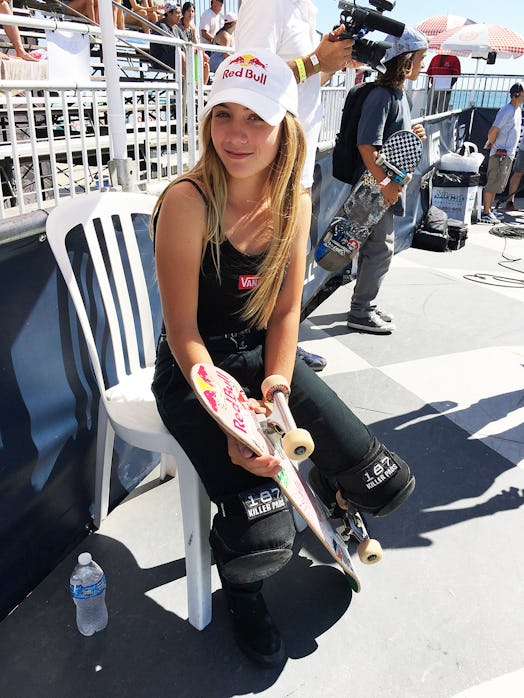 Brighton Zeuner sits in a plastic chair in the shade in front of a set of bleachers, holding a skate...