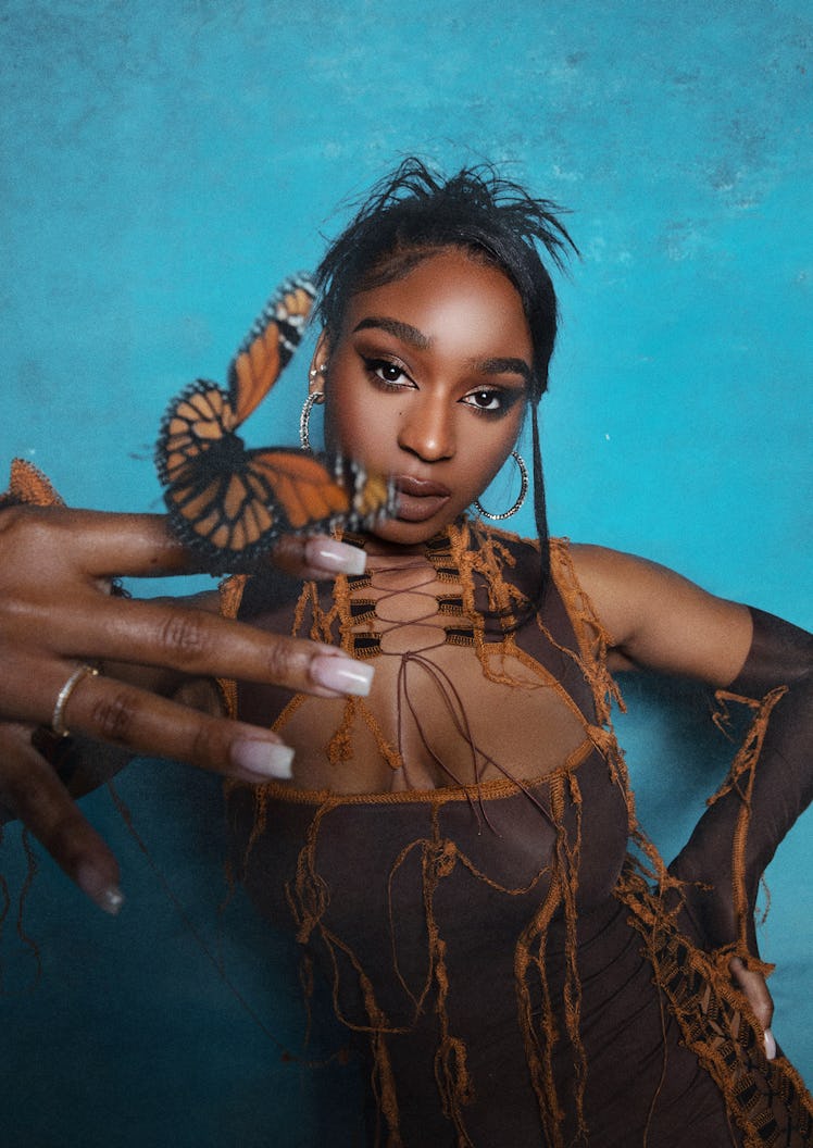 Normani wearing a brown dress and hoop earrings while holding a butterfly on her index finger