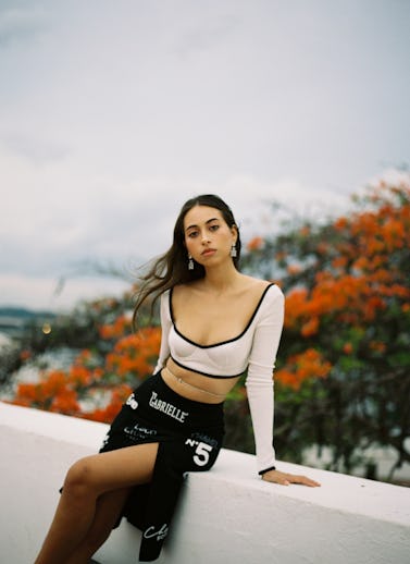 A portrait of Sofía Valdés. She's wearing nude crop top and black skirt while perched on the side of...