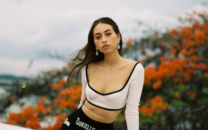 A portrait of Sofía Valdés. She's wearing nude crop top and black skirt while perched on the side of...