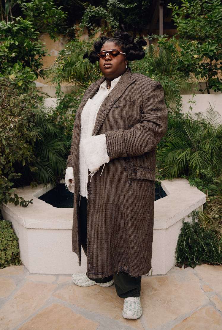 Chika in a brown coat, black sweatpants, white shirt and brown glasses